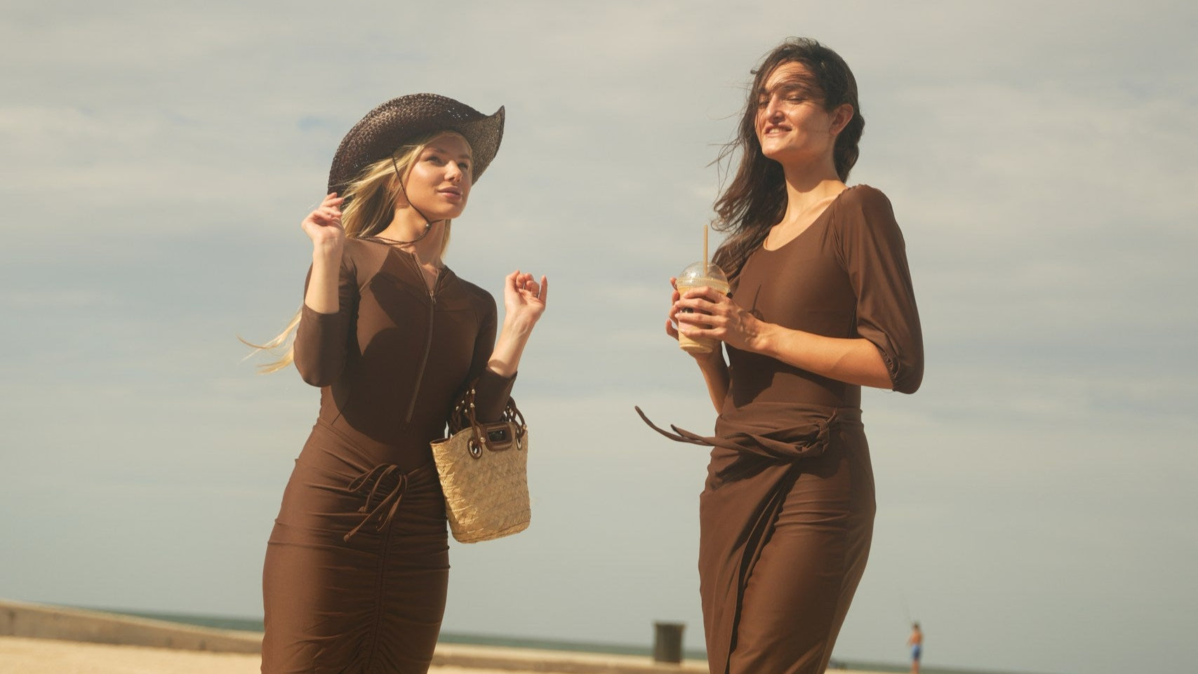 Two women in brown dresses standing on a sandy beach with a clear sky.