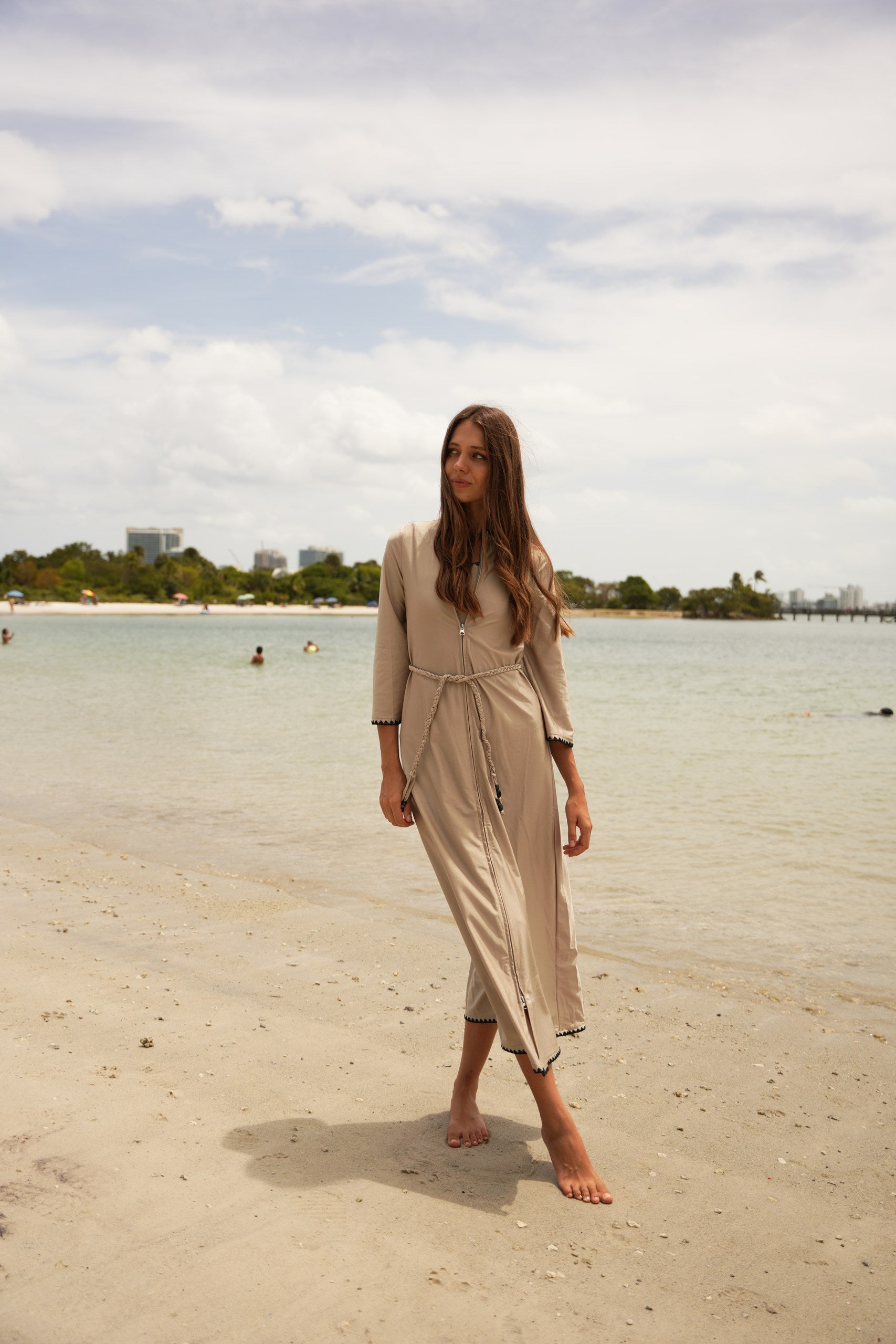 Woman in a beige dress walking on a sandy beach with water and sky in the background