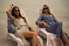 Two women sitting on beach chairs by the water, wearing sunglasses and casual beach attire.